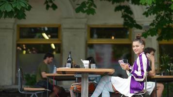 Woman sits at outdoor cafe using smart phone holding a mug and smiles at the camera video