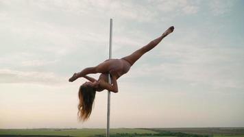 Beautiful athletic woman performs tricks on the pole against the background of sunset in the field. Beauty and body care. Female sports and fitness. Slow motion