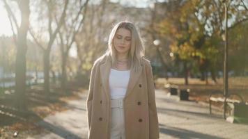 Portrait, Beautiful Business Woman Looks and Smiles at the Camera while Standing in an Autumn City Park on a Bright Sunny Day. Attractive Blonde in a Brown Coat. Positive Female Emotions. Slow Motion video