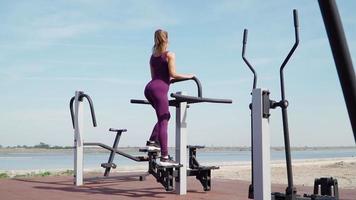 smal atletisk kvinna i en sporter lila träningsoverall övning på en stationär cykel på träna jord i en stad parkera. kondition utomhus. långsam rörelse video