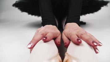 la ballerine est assise sur un fond blanc et passe ses mains sur le tutu noir et les jambes en chaussons de pointe. ballet. gros plan au ralenti video