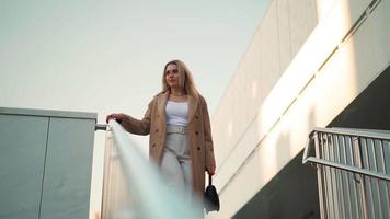 atractiva mujer de negocios con abrigo largo marrón y zapatos blancos de tacón alto camina por las escaleras con la ciudad al fondo. hermosa rubia camina por la ciudad de otoño. camara lenta. video