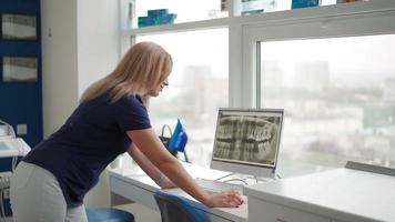Beautiful Woman Dentist Examines a Picture of a Human Jaw on a Computer Monitor Screen. Woman Professional Orthodontist Smiles at the Camera. Concept of Dental Treatment and Medicine. Slow Motion