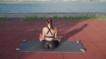 atletisch vrouw zittend in lotus positie en aan het doen yoga Aan meer achtergrond. mooi meisje oefenen buitenshuis. uitrekken opdrachten en flexibiliteit. sport en gezond levensstijl concept. langzaam beweging video