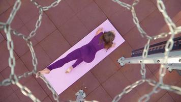 Young athletic woman in a sports purple tracksuit does exercise for stretching. Fitness outdoors. Slow motion. Top view through a basketball hoop video