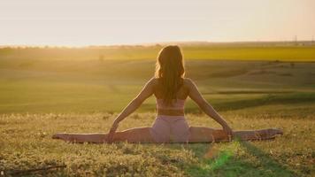 vrouw is beoefenen yoga en doet splitst buitenshuis Aan achtergrond mooi veld- en zonsondergang. geschiktheid in natuur. uitrekken Gezondheid welzijn. sport motivatie en vrijheid. langzaam beweging video