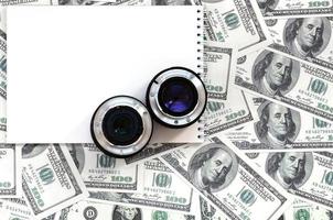 Two photographic lenses and white notebook lie on the background of a lot of dollar bills. Space for text photo