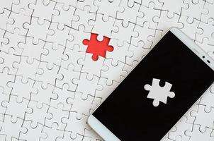 A modern big smartphone with several puzzle elements on the touch screen lies on a white jigsaw puzzle in an assembled state with missing elements photo
