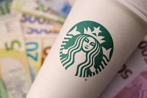 KHARKIV, UKRAINE - DECEMBER 16, 2021 White paper cup with Starbucks logo and money bills. Starbucks is the world's largest coffee house with over 20,000 stores. photo