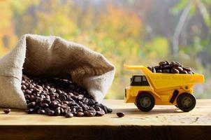 A small yellow toy truck is loaded with brown coffee beans around a full bag of grains. A car on a wooden surface against a background of autumn forest. Extraction and transportation of coffee photo