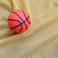 Small orange rubber basketball lies on a yellow sport jersey clothing fabric texture and background with many folds photo