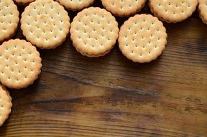 A round sandwich cookie with coconut filling lies in large quantities on a brown wooden surface. Photo of edible treats on a wooden background with copy space