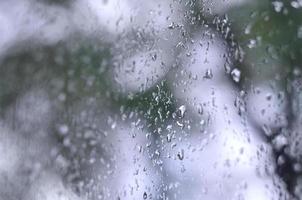 A photo of rain drops on the window glass with a blurred view of the blossoming green trees. Abstract image showing cloudy and rainy weather conditions