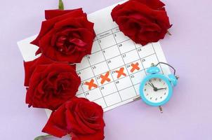 Blue alarm clock and red rose flowers on menstrual period calendar with red cross marks photo