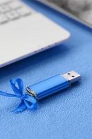 Brilliant blue usb flash memory card with a blue bow lies on a blanket of soft and furry light blue fleece fabric beside to a white laptop and smartphone. Classic female gift design for a memory card photo