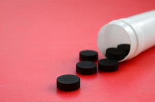 Several black tablets fall out of the plastic jar on the red surface. Background image on medical and pharmaceutical topics. Activated Charcoal photo