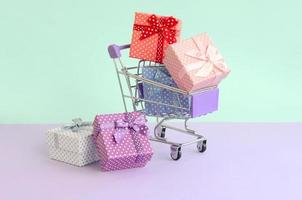 Small gift boxes of different colors with ribbons lies in shopping cart on a violet and blue pastel background photo