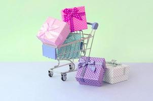 pequeñas cajas de regalo de diferentes colores con cintas en carrito de compras sobre un fondo violeta y azul pastel foto