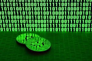 A pair of bitcoins lies on a cardboard surface on the background of a monitor depicting a binary code of bright green zeros and one units on a black background. Low key lighting photo