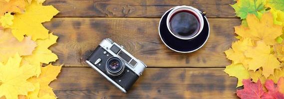 A cup of tea and an old camera among a set of yellowing fallen autumn leaves on a background surface of natural wooden boards of dark brown color photo