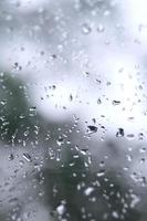 A photo of rain drops on the window glass with a blurred view of the blossoming green trees. Abstract image showing cloudy and rainy weather conditions