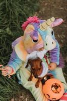 little laughing girl and a dwarf dachshund in Halloween costumes with a pumpkin basket for sweets outdoors. a girl in a rainbow unicorn kigurumi costume, a dog in a dress with a full skirt. top view photo