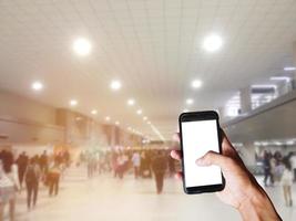 mano sosteniendo un smartphone negro con pantalla en blanco detrás de muchos turistas que se mueven dentro del aeropuerto con un fondo borroso. espacio para texto o diseño foto