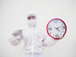 retrato de médico o científico con uniforme de suite ppe sosteniendo un despertador rojo y mirando la cámara con varios gestos. covid-19 concepto aislado fondo blanco foto