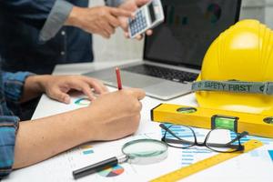 Asian engineer and partner team learn and talk with house plan project blueprint, graph and yellow helmet and engineering construction tools on table work. photo