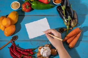 Vegetables are laid out around a sheet of paper and a pencil. Empty space for text. Vegetables, empty blank for recipe on a blue background. photo