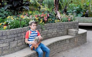 Portrait kid holding plush toy sitting in the garden,Happy boy looking out with thinking face,Child wearing cap hat, carrying backpack enjoying time in the park,Children exploring with nature concept photo