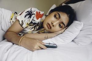 Sad young woman frowning from the bed waiting for a call while holding the smartphone. photo