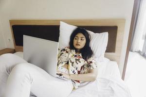 Serious concentrated young woman working from the bed. Hold the laptop on the knees and type on the keyboard. photo