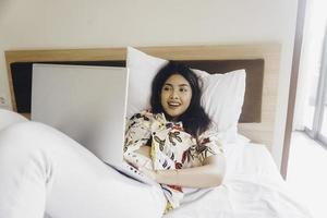 Happy young woman working from the bed. Hold the laptop on the knees and type on the keyboard. photo