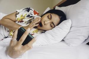 A sleepy young woman yawning while holding phone on the bed. photo