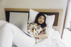 Happy young woman working from the bed. Hold the laptop on the knees and type on the keyboard. photo