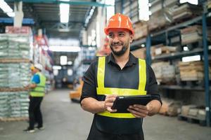 Professional manager man employee smile using tablet check stock working at warehouse. Worker wearing high visibility clothing and a hard hat, helmet checking and count up goods or boxes for delivery photo