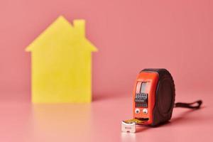 House renovation funny concept. Metal tape measure and other repair items. Home repair and redecorated concept. Yellow house shaped figure on pink background. photo