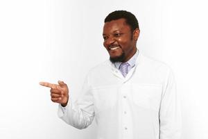 Happy black doctor man with small beard in white coat points to right copy space on white background photo