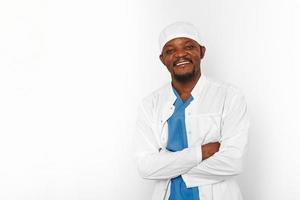 sonriente médico cirujano negro barbudo en bata blanca y gorra con brazos cruzados aislado en blanco foto