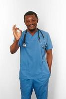 Smiling black surgeon doctor bearded man in blue coat with stethoscope makes OK gesture photo