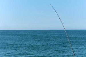 Fishing rod against blue ocean or sea background, copy space. Waiting for biggest haul. Meditative relax sport. photo