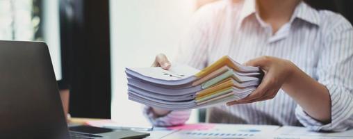 Business Documents concept  Employee woman hands working in Stacks paper files for searching and checking unfinished document achieves on folders papers at busy work desk office. Soft focus photo