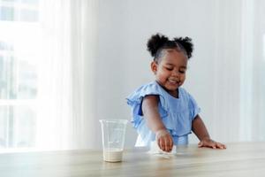 una niña linda disfruta limpiando la leche derramada sobre la mesa. centrarse en un vaso de leche. foto