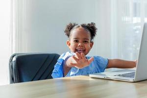 little girl of color Wear a blue shirt, sit at a wooden table, play computer games online. happily internet connection concept photo