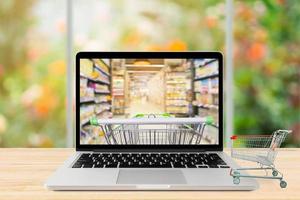 laptop computer and shopping cart on wood table with window and garden abstract blur background grocery online concept photo