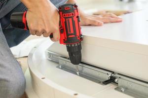 man assembling white table furniture at home using cordless screwdriver photo