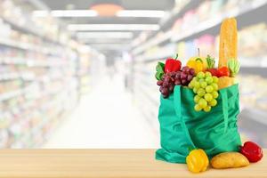 Fresh fruits and vegetables in reusable green shopping bag on wood table top with supermarket grocery store blurred defocused background with bokeh light photo