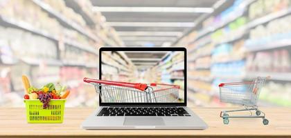 supermarket aisle blurred background with laptop computer and shopping cart on wood table grocery online concept photo