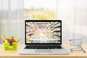 laptop computer and shopping cart on wood table with window curtain abstract blur background grocery online concept photo